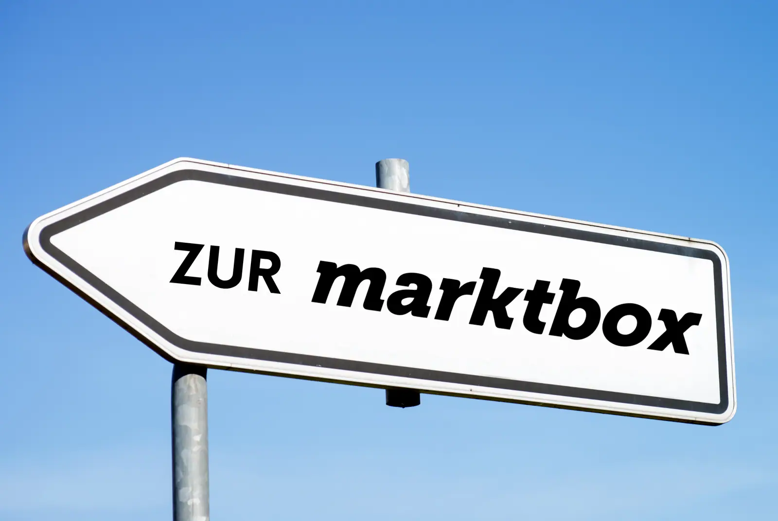 Weißes Hinweisschild in Pfeilform mit der Aufschrift „ZUR marktbox“ zeigt vor blauem Himmel nach links