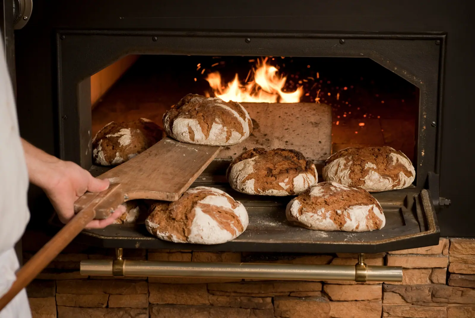 Bäcker entnimmt frisch gebackene, rustikale Brotlaibe mit einem Holzschieber aus einem lodernden Steinofen