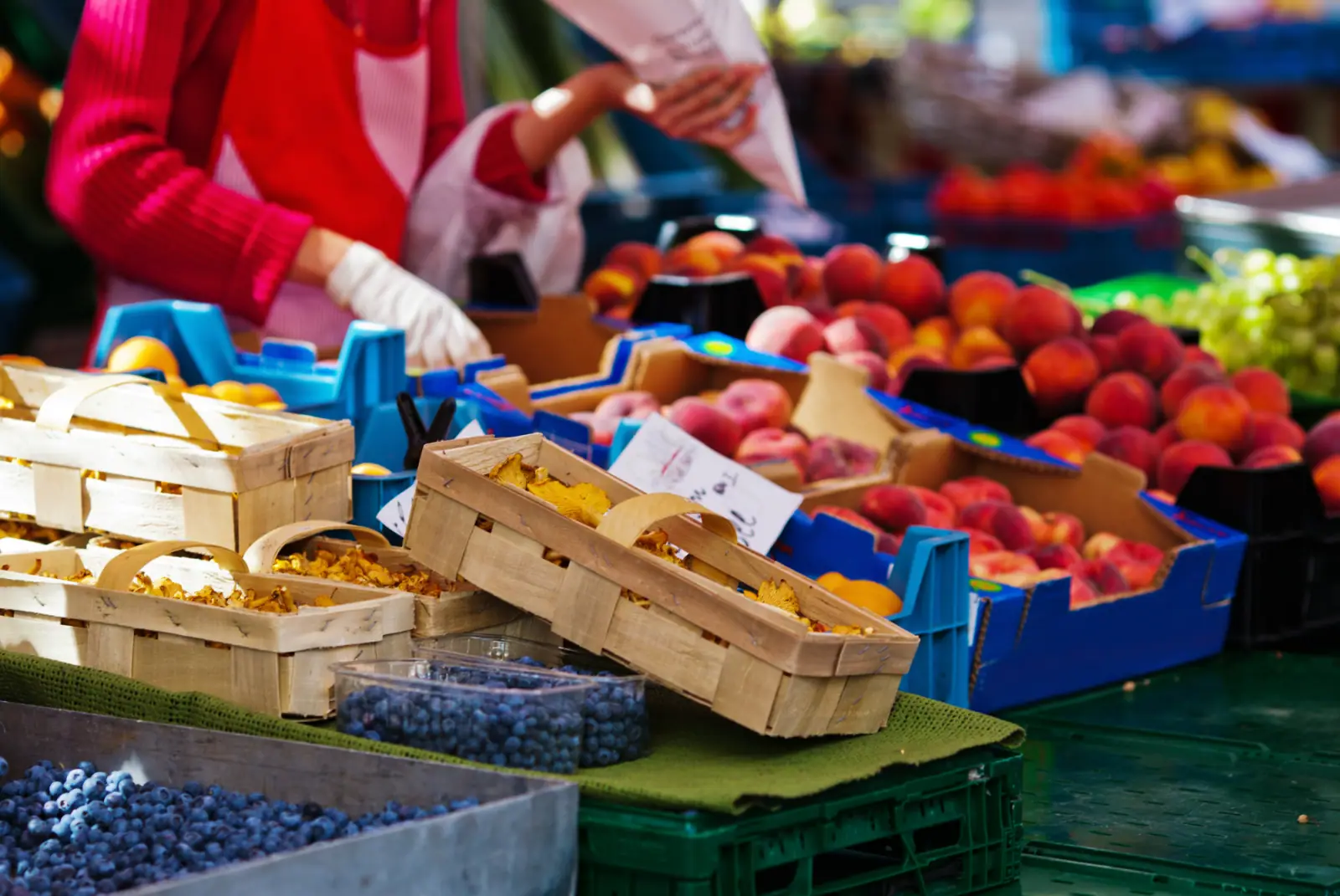 Marktstand mit Pfifferlingen und Obst sowie einer Person, die mit Handschuhen Lebensmittel verpackt