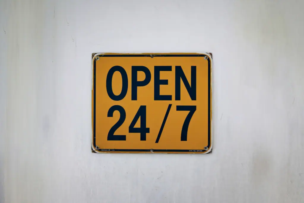 Gelbes quadratisches Schild mit der schwarzen Aufschrift OPEN 24/7 an einer weißen Wand