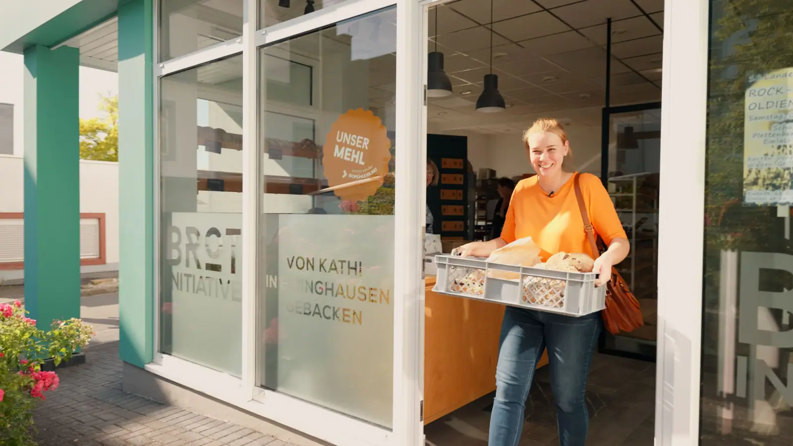 Lächelnde Frau trägt eine Kiste mit frischem Brot aus dem Eingang einer Bäckerei