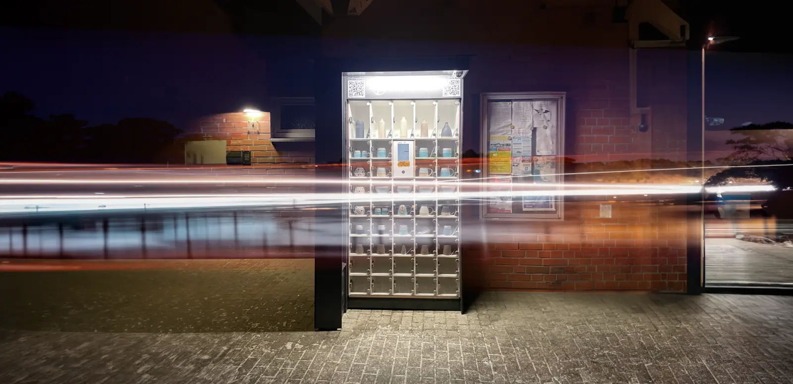 Beleuchtete marktbox mit Keramik in Fächern vor einer Backsteinwand bei Nacht mit dynamischen Lichtspuren