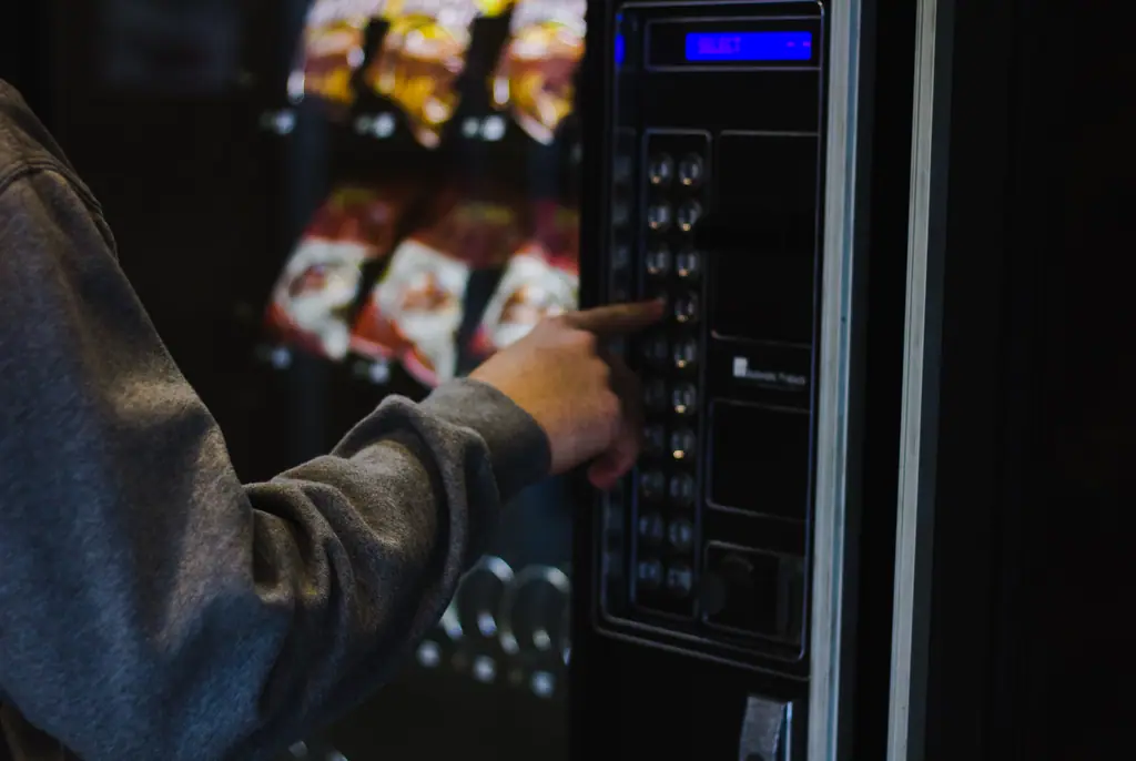 Eine Hand bedient das Tastenfeld eines Verkaufsautomaten mit Snacks im Hintergrund