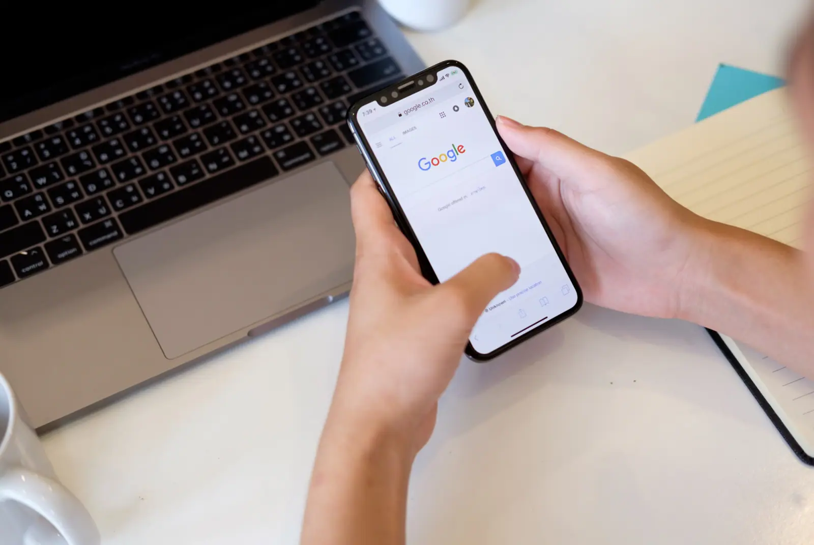 Smartphone mit der Google-Suche in den Händen einer Person neben einem Laptop auf einem weißen Tisch