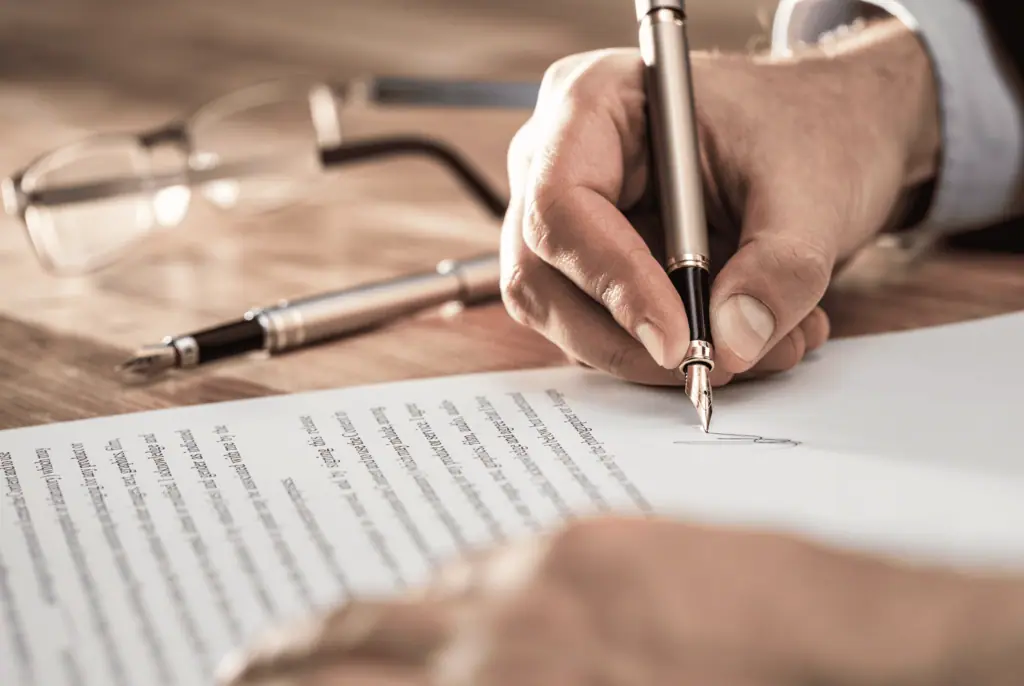 Hand setzt mit einem Füllfederhalter eine Unterschrift unter ein Dokument auf einem Schreibtisch