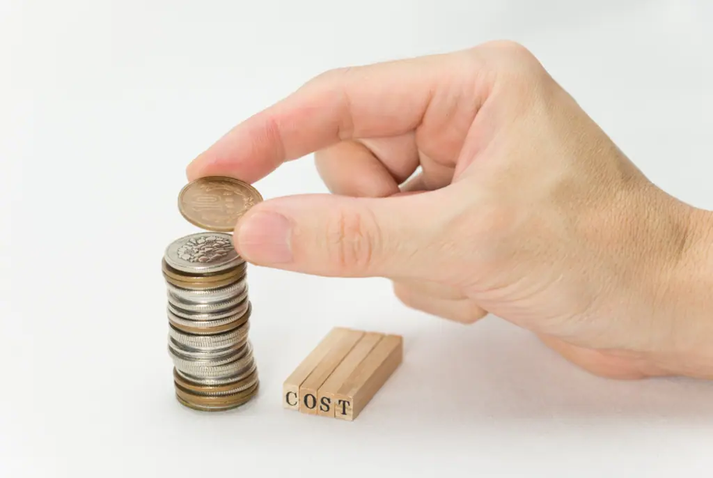 Hand legt eine Münze auf einen Geldstapel neben Holzplättchen mit der Aufschrift COST