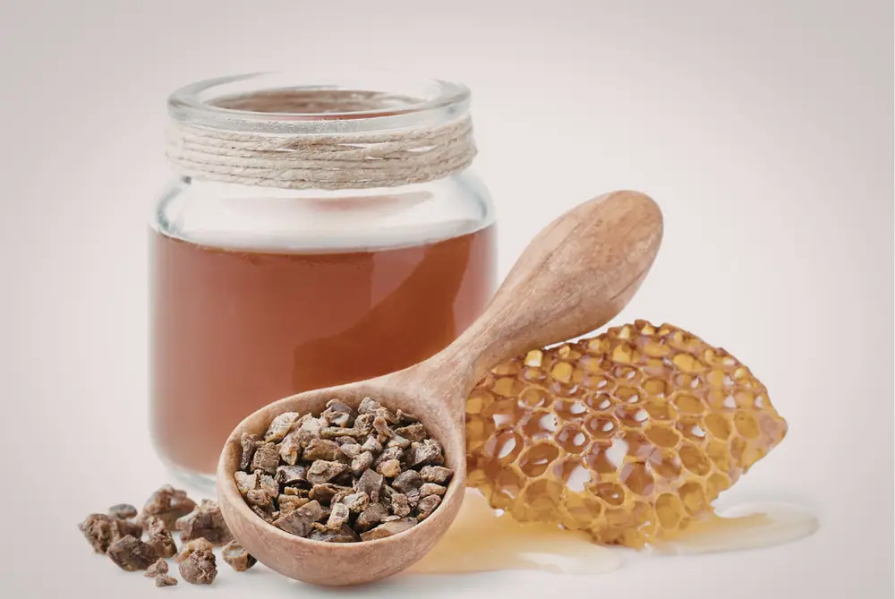 Honigglas neben einer Honigwabe und einem Holzlöffel mit Bienenprodukten auf hellem Untergrund