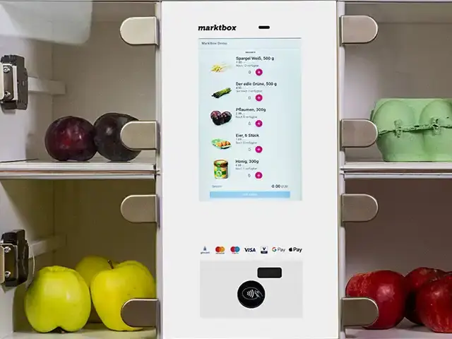 Marktbox-Terminal mit Display zur Artikelauswahl und Kartenzahlungssymbolen zwischen Regalen mit Obst und Eiern