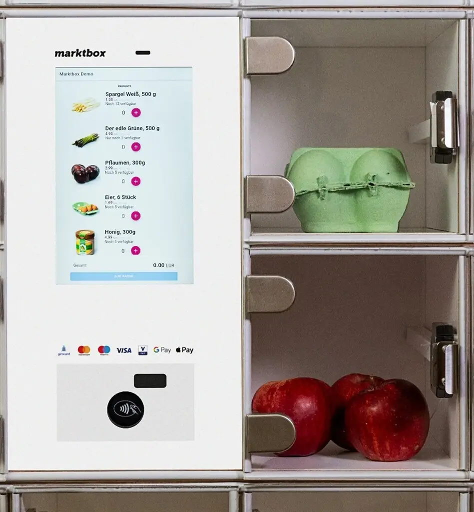 Digitale Marktbox mit Touchscreen und Zahlterminal neben Fächern mit einem Eierkarton und roten Äpfeln