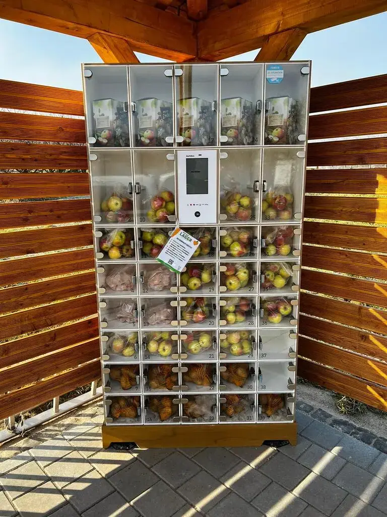 Digitaler marktbox-Verkaufsautomat mit Glasfächern voller Äpfel und Fruchtsäften unter einem Holzdach