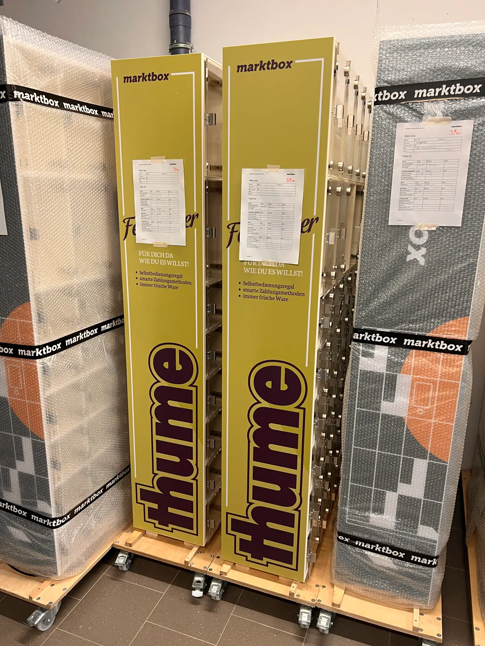 Gelbe Marktbox-Automaten mit thume-Aufdruck und verpackte Module auf Rollwagen in einer Produktionshalle