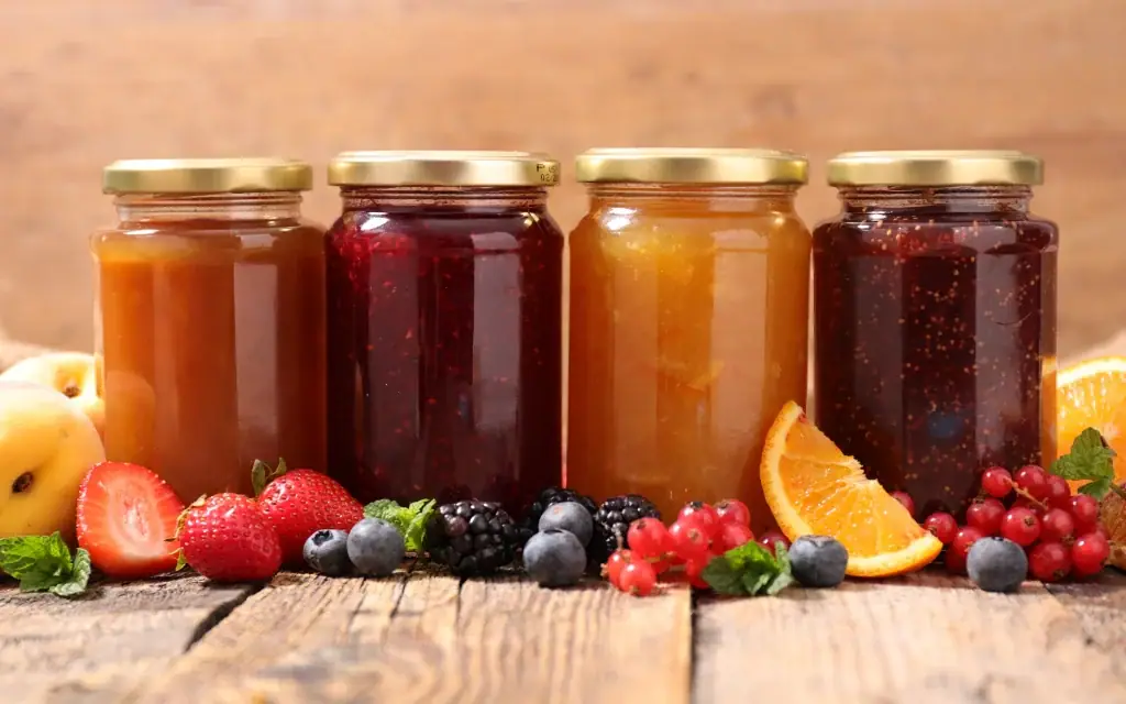 Vier Gläser mit verschiedenen Marmeladen und frischen Früchten auf einem rustikalen Holztisch